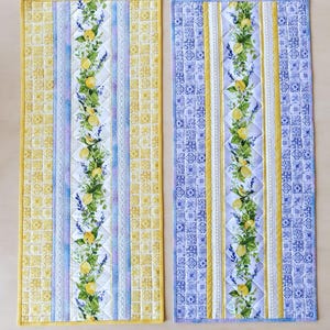 May include: Two rectangular quilted table runners. One is yellow and blue with a lemon and lavender floral design down the centre. The other is blue and white with the same floral design. Both have a yellow border.
