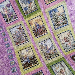 May include: A quilted blanket featuring rectangular panels with illustrations of fairies among flowers. The panels are framed in green and blue, set against a lavender and pink background. The quilt has a floral theme, with the text "Flower Fairies" and "NovaQuilt on Etsy".