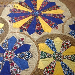 May include: Circular placemats with a Ukrainian floral design. The placemats feature a central flower motif with blue, yellow, and patterned fabric accents. The placemats have a woven, textured border. The text "Greatness of Ukraine NovaQuilt on Etsy" is visible.