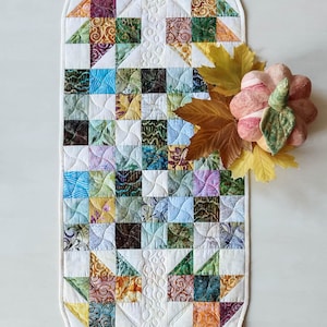 Puede incluir: Un camino de mesa acolchado con un diseño de patchwork con cuadrados y triángulos en varios colores como verde, naranja, azul y morado. El camino de mesa tiene un borde blanco y costuras decorativas. Una calabaza decorativa y hojas se colocan en el lateral.