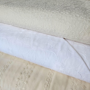 May include: Close-up of three rolls of fabric in varying shades of white and cream. The top roll features a small floral pattern, the middle roll has a larger floral design, and the bottom roll has a striped pattern with floral accents. The fabrics appear to be cotton or linen.