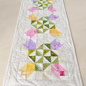 May include: A handmade, quilted table runner featuring a floral design. The runner showcases a repeating pattern of colorful tulip-like shapes in shades of yellow, pink, and purple, set against a white background. The runner is approximately 36 inches long.