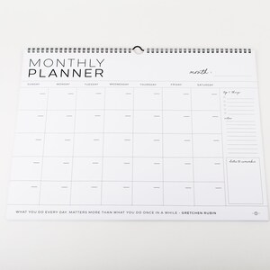 May include: A white monthly planner with a spiral binding and a hanging loop. The planner features a grid layout for each day of the week, with space for notes and a to-do list. The text "MONTHLY PLANNER" is at the top.