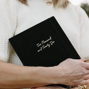 May include: A black journal with the text "Two Thousand and Twenty Six" in gold script. The journal is held by a person wearing a white sweater and gold bracelet. The background is blurred.
