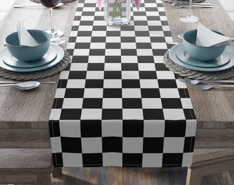 May include: A black and white checkered table runner is displayed on a wooden table, set for a meal. The runner is the focal point, with a repeating pattern of black and white squares. The table is set with plates, bowls, silverware, and wine glasses.