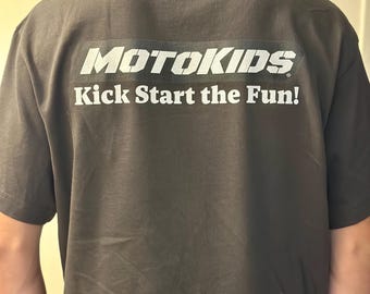May include: Dark gray t-shirt with the text "MOTOKIDS" in white block letters above the phrase "Kick Start the Fun!" in white. The shirt is a short-sleeved crew neck.
