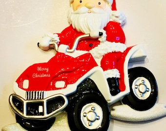 May include: A red and white ATV ornament with a Santa Claus figure driving it. The ornament has a glittery white base and the text "Merry Christmas" is written on the side.