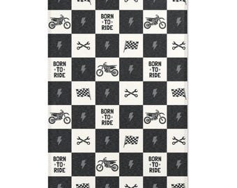 May include: A black and white checkered rug featuring motorcycle, lightning bolt, wrench, and checkered flag graphics. The phrase "BORN + TO + RIDE" is repeated throughout the design. The rug has a textured appearance.