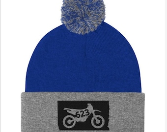 May include: A blue and gray knit beanie with a pom pom on top. The beanie has a black silhouette of a dirt bike with the number 623 on it.
