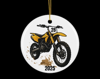 May include: A white ceramic ornament featuring a yellow and black dirt bike illustration. The bike has the number 28 on the front and the year 2025 below. The ornament hangs from a gold string.