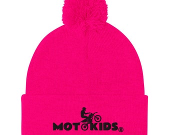 May include: A bright pink beanie with a fluffy pom pom on top. The beanie has a black embroidered logo that says "MOTO KIDS" with a silhouette of a person riding a dirt bike.