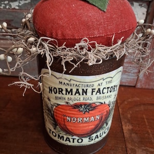 May include: A decorative tomato-shaped pin cushion made of red fabric, with a green leaf accent. It sits atop a vintage-style can labeled "Norman Tomato Sauce." The can is brown with a cream-colored label and a tomato illustration.