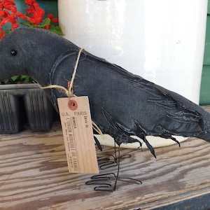 May include: A black fabric crow with a wire stand. The crow has a distressed look and is decorated with stitching. A brown paper tag is attached to the crow with twine. The tag reads "U.A. PARIS, ETO 44, BERNELIER, L'UNION AGRICOLE DE FRANCE".
