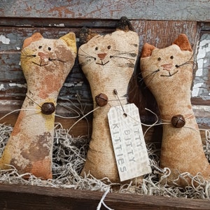 May include: Three handmade fabric cat figurines with brown, orange, and yellow tones. The cats are standing upright and have a tag that reads "itty bitty kitties".