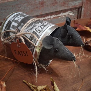 May include: Two black fabric rat figures emerge from a repurposed tin can. The can is wrapped with twine and has a pumpkin-shaped tag that reads "RATS!". The can has a black and white label. The rats have long whiskers.