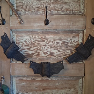May include: A decorative garland featuring three black fabric bats suspended on a thin string. Each bat has a textured surface, small dark eyes, and outstretched wings. The bats are arranged in a row, connected by a light-colored cord.