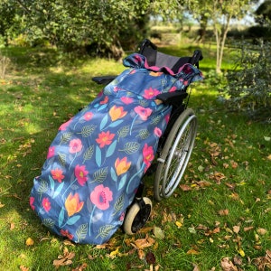 Op de afbeelding: Een rolstoelcover met een bloemenpatroon. De cover is marineblauw met roze, oranje en gele bloemen en groene bladeren. De cover is over een rolstoel op een grasveld gedrapeerd.