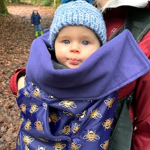 Könnte beinhalten: Ein Baby trägt eine blaue und goldfarbene Babytrage mit einem blauen Fleecefutter. Das Baby trägt eine blaue Strickmütze und hat einen neugierigen Gesichtsausdruck.