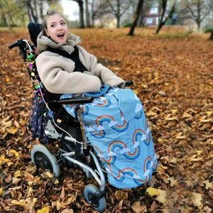 Könnte beinhalten: Ein Rollstuhl mit einer hellblauen Decke mit einem Regenbogen- und Wolkenmuster. Der Rollstuhl hat einen silbernen Rahmen und schwarze Räder. Der Hintergrund ist ein Park mit Herbstlaub.