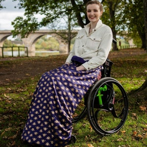 Può includere: Una persona con una camicia bianca è seduta su una sedia a rotelle con una coperta a motivi viola e dorati drappeggiata sulle sue ginocchia. La coperta ha un motivo ripetitivo di api. La persona sorride e guarda la telecamera. La sedia a rotelle si trova in un parco con erba verde e un ponte sullo sfondo.