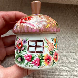 Puede incluir: Un pequeño tarro de cerámica con una tapa en forma de sombrero de seta. La tapa es marrón con detalles florales pintados en blanco y rojo. La base blanca presenta una ventana cuadrada pintada con un marco marrón, rodeada de coloridos motivos florales.
