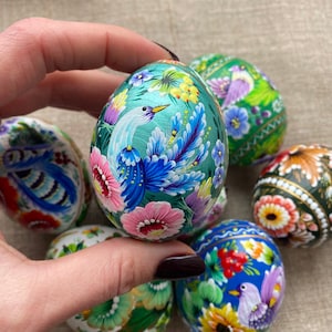 May include: Six hand-painted Easter eggs with floral and bird designs. The eggs are painted in various colours, including green, blue, pink, yellow, and white. The eggs are arranged in a circle on a beige surface.