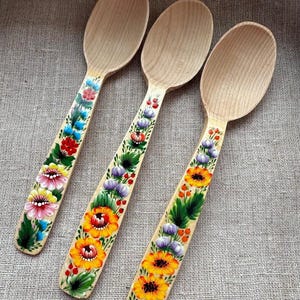 Può includere: Tre cucchiai di legno con disegni floreali dipinti a mano. I manici dei cucchiai presentano fiori colorati nelle tonalità del rosso, giallo, rosa e blu, con foglie verdi. I cucchiai sono disposti su una superficie strutturata di colore neutro.