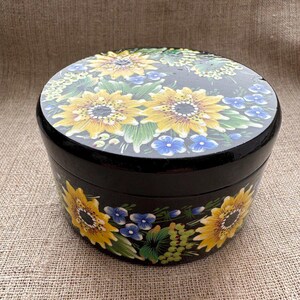 May include: A round, black wooden box with a lid, hand-painted with yellow sunflowers, blue forget-me-nots, and green foliage. The floral design wraps around the sides and top of the box, creating a decorative storage container.
