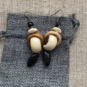 Puede incluir: Un par de pendientes de madera hechos a mano. Cada pendiente presenta una combinación de cuentas de madera beige claro, marrón oscuro y negro. Los pendientes están suspendidos de ganchos de metal oscuro y se exhiben en una bolsa de tela gris.