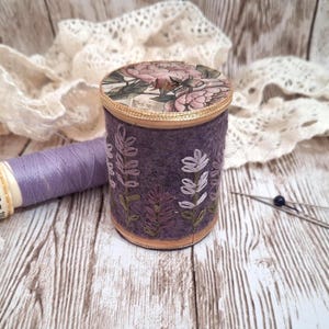 May include: A cylindrical purple felt pincushion featuring embroidered lavender sprigs on the side and a floral patterned top. Beside it lies a spool of light purple thread labelled 100 m and two sewing needles with dark beads on the ends.