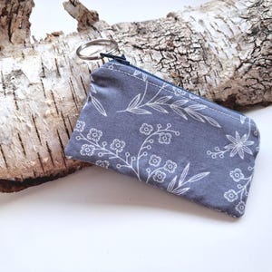 May include: A small, rectangular fabric coin pouch in a muted slate blue color featuring a white line-art pattern of delicate flowers and leaves. The pouch includes a dark blue zipper closure along the top edge and a silver metal split key ring.