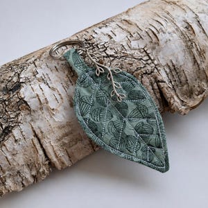 May include: A fabric keychain shaped like a leaf in a muted sage green color, featuring a repeating pattern of dark green acorns. The leaf is quilted with white stitching and includes a silver-toned metal keyring and a small silver branch charm.