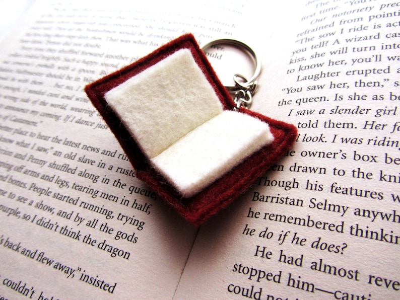 May include: A red felt book keychain with white pages, open and ready for a story. The keychain is attached to a silver key ring.