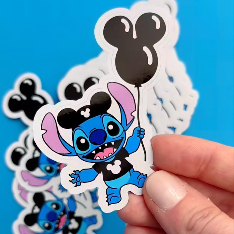 Puede incluir: Un personaje azul Stitch que lleva una camisa negra con un logotipo de Mickey Mouse y orejas de Mickey Mouse. Stitch sostiene un globo negro de Mickey Mouse.
