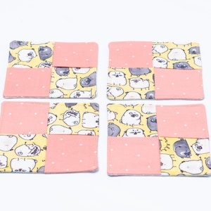 May include: A set of four square fabric coasters featuring a patchwork design. Each coaster alternates between a light pink fabric with small white dots and a yellow fabric printed with a repeating pattern of cartoon pugs in white and gray.