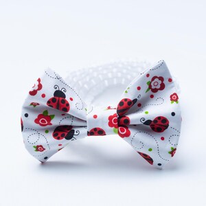 May include: A white bow tie featuring a repeating pattern of red ladybugs, small red flowers with green leaves, and black dots. The fabric has thin, swirling black lines connecting the elements, and a white elastic band is attached to the back.