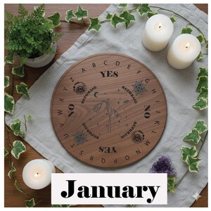 May include: A wooden Ouija board with engraved letters, numbers, and symbols. The board features the words "YES," "NO," and "REPHRASE." Two lit candles and a small potted plant are visible. The word "January" is displayed at the bottom.