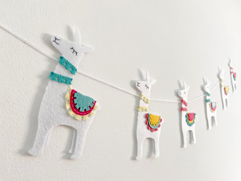 May include: A white felt llama garland featuring five individual llama cutouts strung together on a thin white cord. Each llama has embroidered closed eyes and a mouth, with colorful felt saddles and neck trim in teal, yellow, and pink.