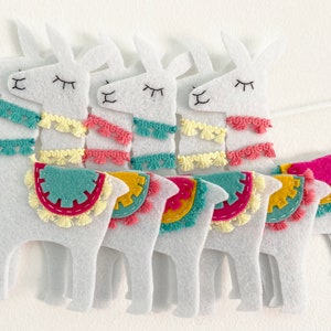 May include: A garland featuring five white felt llama cutouts strung together. Each llama has embroidered closed eyes and a mouth, decorated with colorful fringe trim in teal, yellow, and pink, and a saddle blanket in matching felt colors.