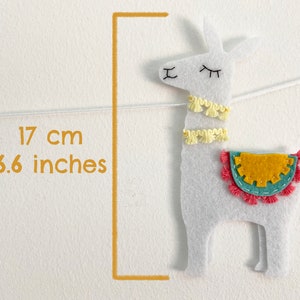 May include: A white felt llama ornament featuring two yellow tassel collars, a teal and mustard yellow saddle with pink fringe, and embroidered facial features. A graphic indicates a height of 6.6 inches, suspended by a thin white string.