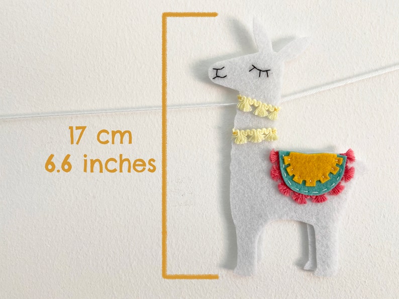May include: A white felt llama ornament featuring two yellow tassel collars, a teal and mustard yellow saddle with pink fringe, and embroidered facial features. A graphic indicates a height of 6.6 inches, suspended by a thin white string.