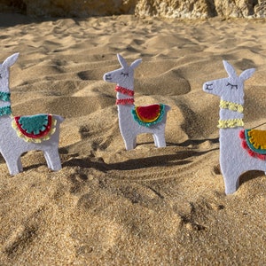 May include: Three white felt llama ornaments with embroidered closed eyes and mouths. Each llama features a colorful saddle blanket with fringe and a matching neck trim in teal, coral, or yellow, standing upright in a row on a sandy surface.