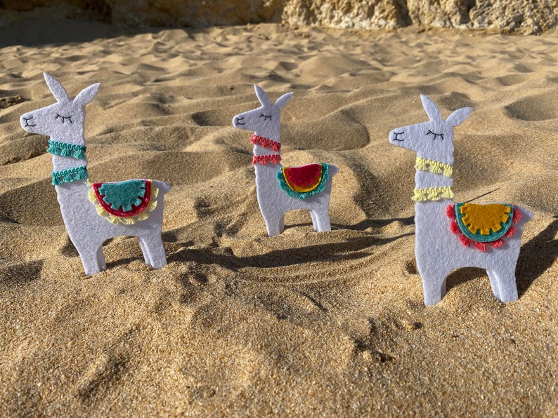 May include: Three white felt llama ornaments with embroidered closed eyes and mouths. Each llama features a colorful saddle blanket with fringe and a matching neck trim in teal, coral, or yellow, standing upright in a row on a sandy surface.