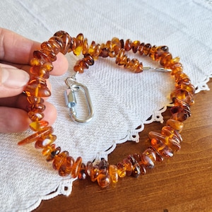 Puede incluir: Un collar hecho de cuentas de ámbar con un cierre plateado. Las cuentas son de un color miel cálido y tienen una forma irregular.