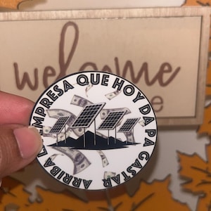 May include: A white round sticker with black text that reads "La Empresa Que Hoy Da Para Gastar Arriba". The sticker features a design of three solar panels with dollar bills flying around them.