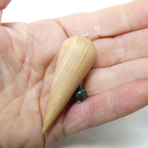 Peut inclure: Un pendule en bois avec une pierre vert foncé au bas. Le pendule a la forme d'une larme et est tenu dans une main.