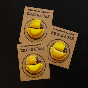 May include: Three brown card stock tags with the text "HANDMADE MAGNET EMPANADAS" printed on them. Each tag features a yellow and brown clay model of an empanada.