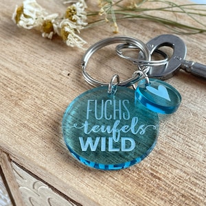 以下が含まれることがあります： 青いアクリル製のキーホルダーで、「FUCHS teufels WILD」という文字と小さな青いハートのチャームが付いています。キーホルダーは銀色のキーリングに取り付けられています。