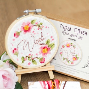 May include: A cross stitch kit with a pink floral design and the text "Weeb Trash" in a cursive font. The kit includes a wooden embroidery hoop, Aida fabric, DMC threads, a chart, instructions, and a needle.
