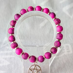 May include: A magenta beaded bracelet with a silver pentagram charm. The bracelet features round, textured beads separated by small silver spacers. The pentagram charm hangs from the centre of the bracelet.